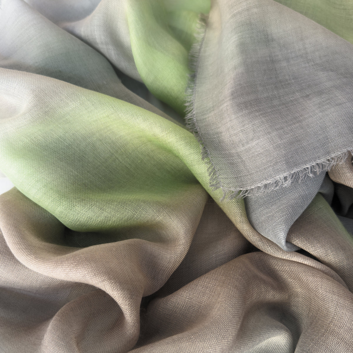 Close-up of a hand-painted Otta wool & silk scarf draped softly, showcasing gentle shades of 
brown, gray and light green. Lightweight artisan accessory with delicate hand-fringed edges, measuring 70 x 197 cm.