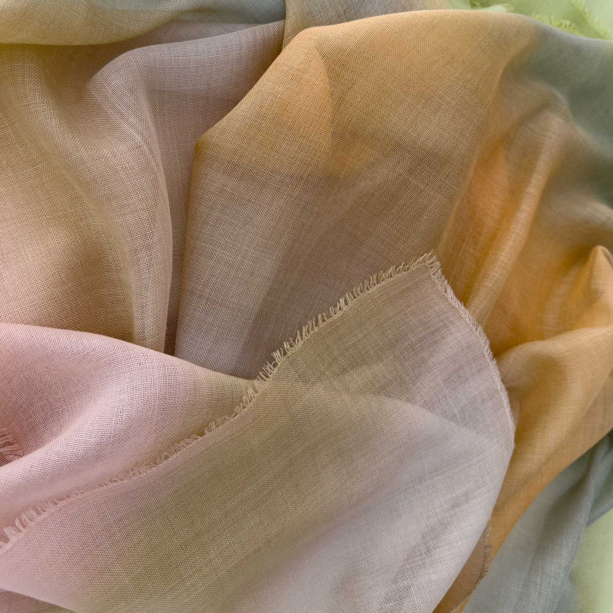 Close-up of hand-painted Otta wool silk blend scarf showcasing soft green, orange and beige shades with delicate hand-fringed and hand-hemmed edges.
