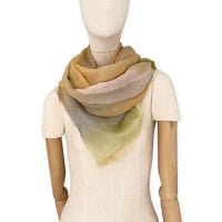 Mannequin wearing an elegant hand-painted Otta wool silk blend scarf on a white background. Warm green, orange and beige tones.
